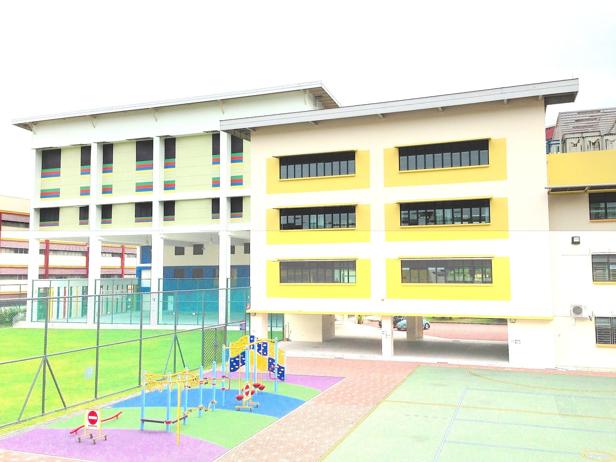 A multi-story school building with yellow accents, colorful playground equipment, a green field, and a sports court.